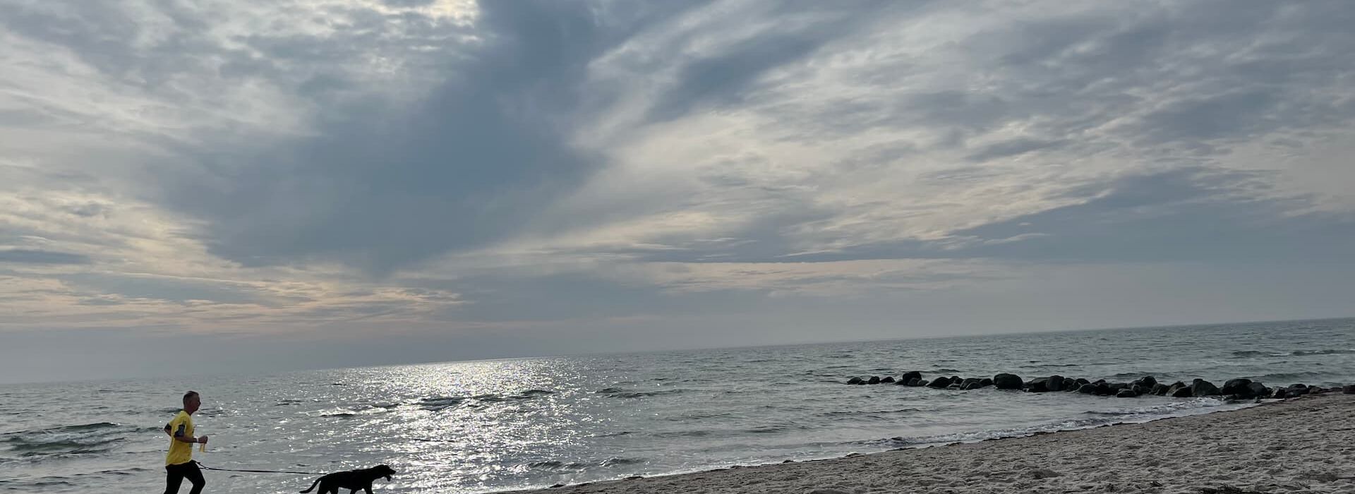 Ein Mann joggt mit seinem Hund am Sandstrand von Grömitz entlang. Die Sonne scheint durch eine dramatische Wolkendecke, das Meer glitzert im Hintergrund. Perfekter Start in den Tag am Hundestrand – Urlaub mit Bewegung und Meerblick.