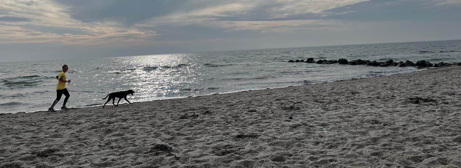 Ein Mann joggt mit seinem Hund am Sandstrand von Grömitz entlang. Die Sonne scheint durch eine dramatische Wolkendecke, das Meer glitzert im Hintergrund. Perfekter Start in den Tag am Hundestrand – Urlaub mit Bewegung und Meerblick.